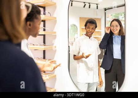 An optician assists a woman in selecting eyewear, offering expert advice. The scene is set in a modern optical store with various glasses displayed. T Stock Photo