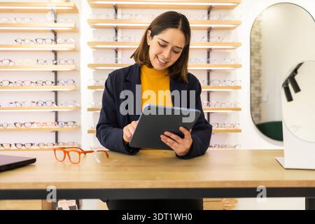 A smiling employee in an optical store holds a tablet while standing behind a counter lined with glasses and frames. Shelves full of eyewear create a Stock Photo