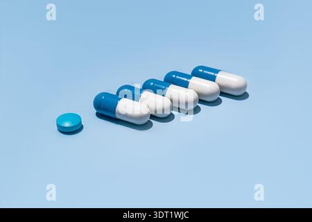 Five blue and white capsules are aligned in a row on a clean blue background, next to a single round blue pill, depicting a medical and pharmaceutical Stock Photo
