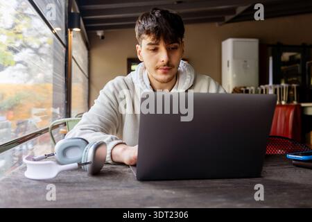 A young man in a hoodie focuses on his work using a laptop, with headphones resting on the desk, illustrating modern technology use in a relaxed indoo Stock Photo