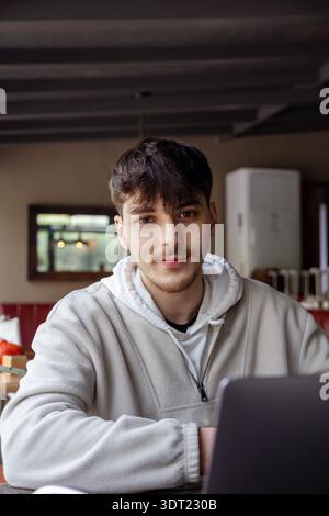 A young man with short dark hair and a light beard sits in a cozy cafe, working on his laptop. He wears a casual grey pullover and a white hoodie, foc Stock Photo