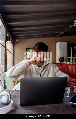 A young man sits in a cozy cafe using a laptop, surrounded by tech gadgets like headphones and a smartphone. He is casually dressed and looks thoughtf Stock Photo