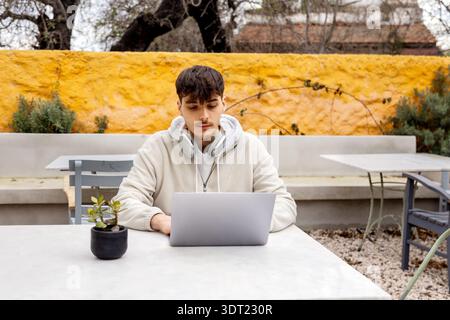 Young man focused on his laptop while working outdoors. Dressed casually with headphones around his neck, he's tapping into technology to complete fre Stock Photo