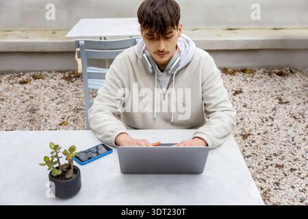A young man working on his laptop outdoors, illustrating a modern freelance lifestyle. He appears engaged in his task, with a smartphone and headphone Stock Photo