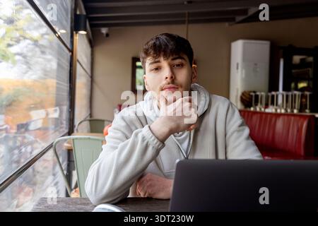 Young man in a light hoodie sitting thoughtfully at a wooden table in a cozy cafe. He is looking at the camera with a contemplative expression, hand o Stock Photo