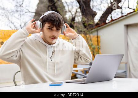 A young man sits outdoors, adjusting his headphones while working freelance on a laptop. He is focused on his technology, embodying a modern tech-savv Stock Photo