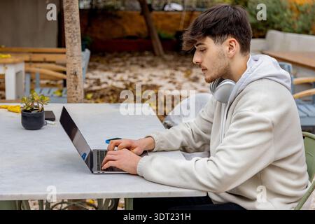 A young man is focused on his laptop, working outdoors as a freelancer. With headphones around his neck, he embodies the modern, tech-savvy lifestyle Stock Photo