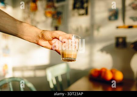 A hand holds a glass of coffee, illuminated by warm sunlight streaming through a window. The cozy indoor setting features a wooden table and softly bl Stock Photo