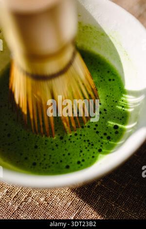 A traditional bamboo whisk gently stirs vibrant, frothy matcha green tea in a white bowl. The scene captures the essence of Japanese tea-making ritual Stock Photo