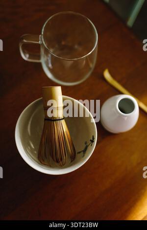 A collection of matcha tea tools including a bamboo whisk, ceramic bowl, and glass cup, artfully arranged on a wooden table, evokes traditional Japane Stock Photo