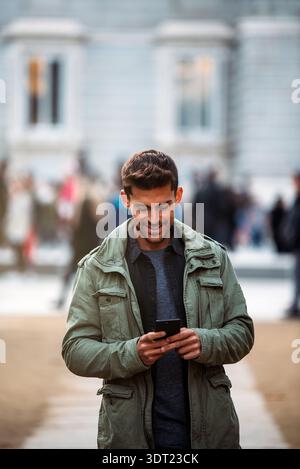 A young man in a casual jacket walks through Madrid while engaged on his smartphone. The city setting and vibrant atmosphere highlight his connectivit Stock Photo