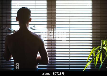 A man stands inside his home, looking out the window through partially opened blinds. Sunlight filters through, illuminating the room and a green plan Stock Photo