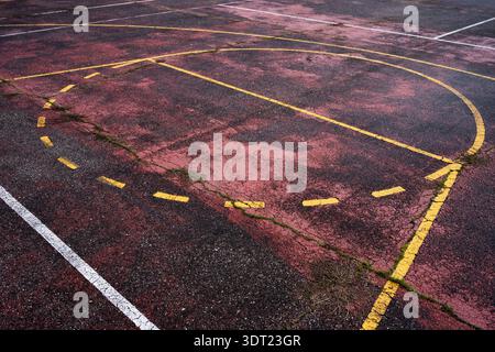 An outdoor basketball court showing signs of age, with faded and cracked yellow and white lines on the asphalt surface. Grass grows through the cracks Stock Photo