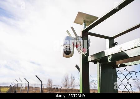 An outdoor surveillance camera is mounted on a metal structure, overseeing a fenced field. The setup enhances security and monitoring capabilities in Stock Photo