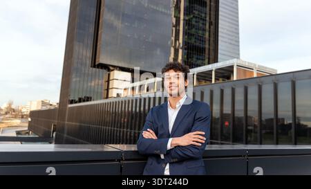 A confident professional in a dark suit stands with arms crossed in an urban business district. The modern glass office building in the background ref Stock Photo