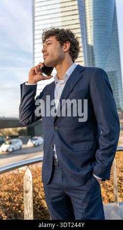 Young professional in a sharp suit making a phone call in a bustling urban environment, with modern skyscrapers in the background. Ideal for business Stock Photo