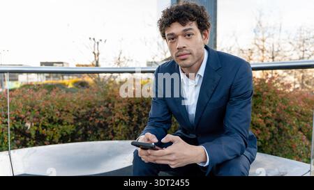 A man in a navy blue business suit sits outdoors, holding a mobile phone and looking at the camera. The background features greenery and urban element Stock Photo