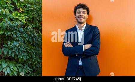 A confident professional in a dark suit stands smiling with arms crossed against a vibrant orange wall, conveying positivity and success. Green foliag Stock Photo