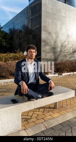 A young businessman in a navy suit sits cross-legged on a concrete bench, wearing white headphones. He relaxes and enjoys the sunny day, with mobile p Stock Photo