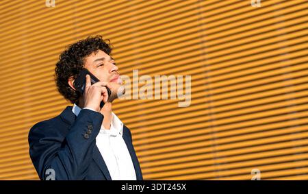 Young professional businessman in a suit making a phone call outside against a striped wall. He looks focused and confident, embracing a clear, sunny Stock Photo