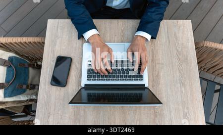 A professional wearing a suit types on a laptop at an outdoor wooden desk. Nearby, a mobile phone and a backpack add to the business-oriented setting. Stock Photo