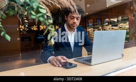 A focused young professional in a suit works on his laptop inside a modern cafe. He is seated at a wooden table, using a mobile phone, surrounded by e Stock Photo