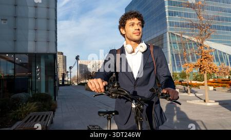 A young business professional in a suit rides a bicycle through a modern urban setting. He carries a backpack and wears headphones, embracing a sustai Stock Photo