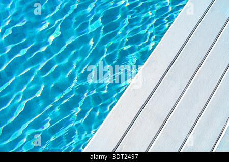 Crystal clear blue water in a swimming pool with sunlight reflecting off the gentle ripples. Adjacent is a pristine white deck, creating a serene and Stock Photo