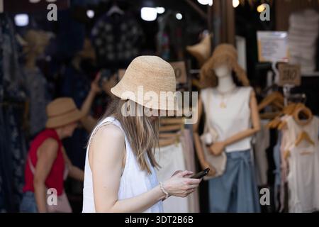 BANGKOK, THAILAND-JAN 25, 2026: Woman in wide-brimmed straw hat using smartphone, shoulder and hand skin texture, side profile, at Chatuchak Market Stock Photo