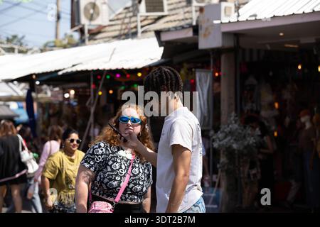 BANGKOK, THAILAND-JAN 25, 2026: Young couple using smartphone in outdoor market, man with dreadlocks, side profile, at Chatuchak Market. Stock Photo