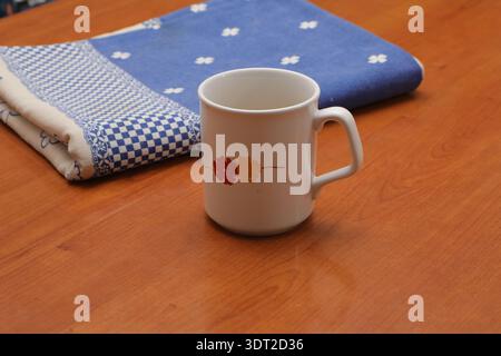 White ceramic mug featuring a red and orange floral design resting on a wooden surface next to a blue patterned cloth Stock Photo