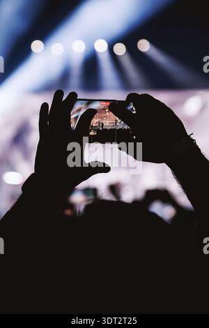 Close-up of hands holding a phone horizontally, showing a clear image of the band on the screen. The stage is lit with cool blue beams and white spotl Stock Photo
