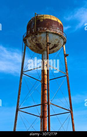 A vertical shot of an old, weathered water tower with peeling paint and visible rust shows signs of age and disuse against a clear blue sky. Stock Photo