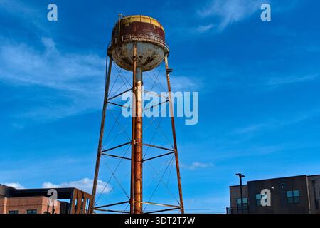 An iconic, weathered water tower with a rusted metal structure and a distressed tank is silhouetted against a bright blue sky dotted with thin clouds Stock Photo