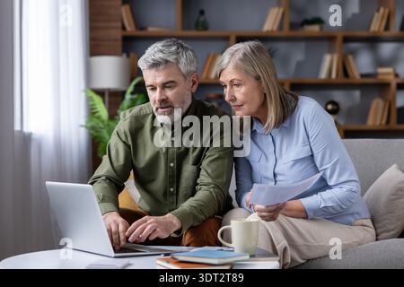 Mature couple on sofa in cozy living room using laptop and papers to review bills, budget and retirement plans, focused on savings, mortgage and investments for future Stock Photo