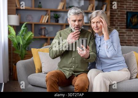 Senior couple sitting on sofa at home, receiving alarming scam email or text message, experiencing shock and frustration from digital literacy challenges and potential financial loss Stock Photo