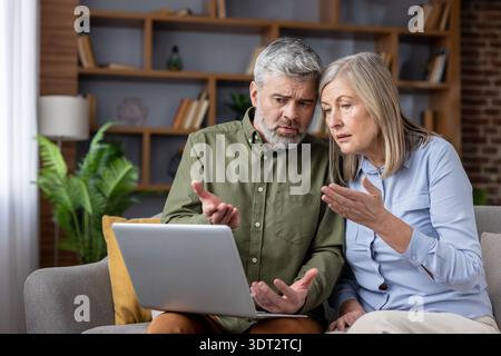 Senior couple sitting on a couch at home feeling frustrated and confused while working with a laptop, experiencing difficulties with technology in a domestic setting Stock Photo