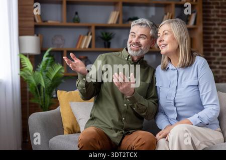 Senior couple sitting closely together on a comfortable sofa in their modern living room, smiling and gesturing, engaging in a pleasant conversation about ideas or plans Stock Photo