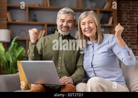 Happy senior couple sitting on sofa at home, smiling at camera and raising their fists in a gesture of triumph while using a laptop for online achievement Stock Photo