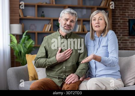 Mature man having a heart attack or medical emergency, clutching his chest in pain while sitting on a sofa next to his worried senior wife at home, seeking help for health symptoms Stock Photo