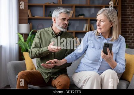 Older man and woman having a serious discussion or conflict at home, with the woman holding a smartphone, representing relationship problems, jealousy, and distrust Stock Photo