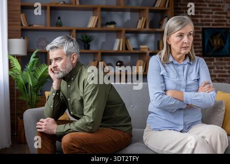 Dissatisfied senior man and woman sitting apart on a sofa at home, feeling upset and distant after a quarrel, showing conflict and communication issues in their marriage Stock Photo