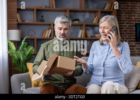 Senior couple on couch arguing with customer service over phone after receiving an empty parcel, frustrated and shocked by an online delivery scam and e-commerce fraud Stock Photo