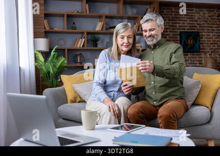 Smiling senior couple reading a letter and holding an envelope in their living room, managing finances and discussing good news with a laptop and documents on the coffee table Stock Photo
