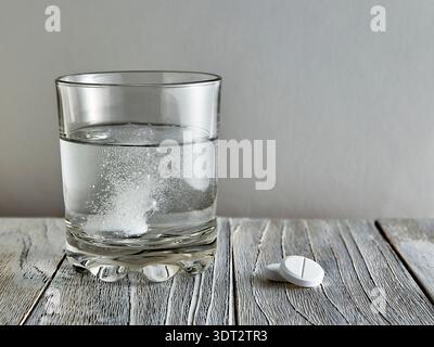 Effervescent tablet in a glass, next to two more tablets on a wooden table Stock Photo