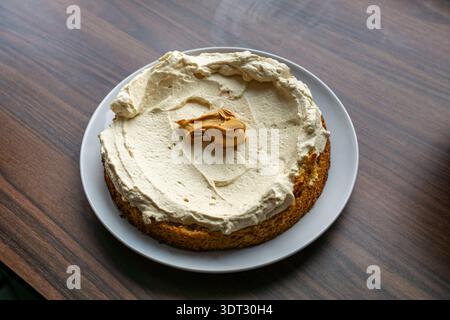 Fresh homemade cake topped with smooth cream frosting and a spoon of peanut butter. Overhead view on a white plate placed on a wooden table. Stock Photo