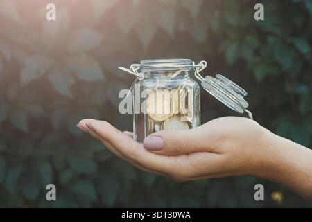 Hand Holding a Glass Jar Filled With Coins Against a Green Background in Daylight Stock Photo