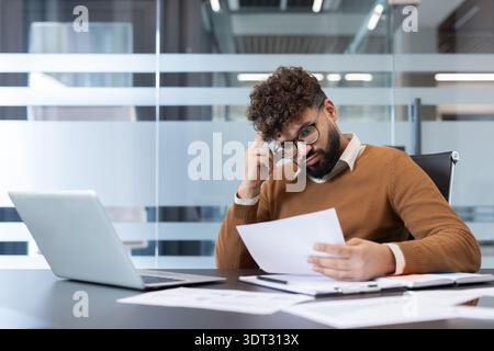 Businessman with glasses and curly hair feeling stressed and overwhelmed while reviewing documents, and working with a laptop at his desk in a contemp Stock Photo