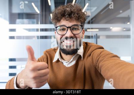 Smiling man with curly hair and beard wearing eyeglasses giving a thumbs up gesture, making a selfie or having a video call from a modern office setti Stock Photo