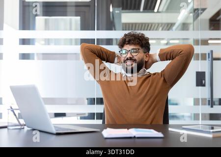 Confident young man smiling, leaning back in his office chair with hands behind head, enjoying a moment of relaxation and success in a modern workspac Stock Photo